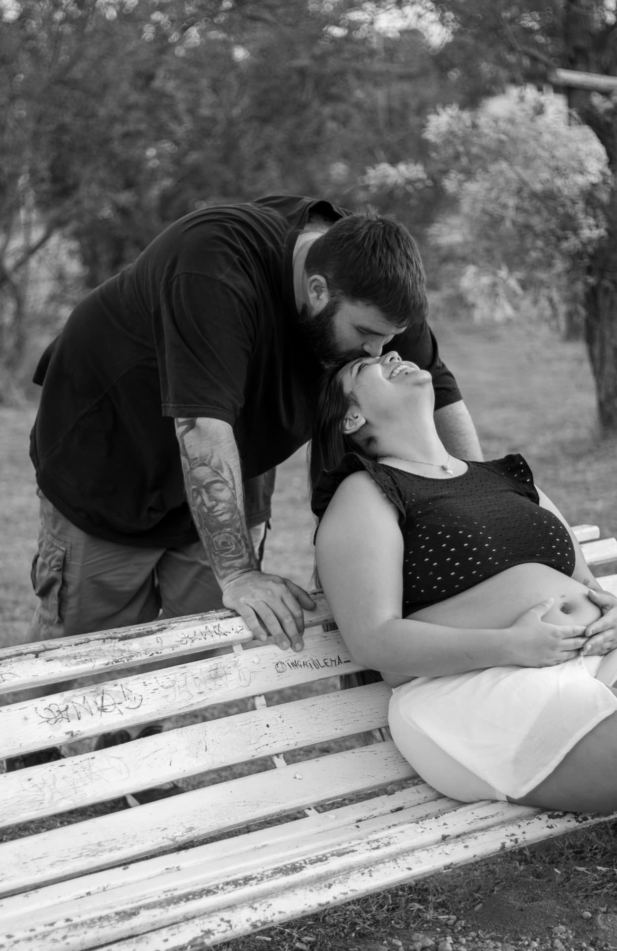 Pareja en una plaza, él besando su frente mientras ella sostiene la panza, en blanco y negro